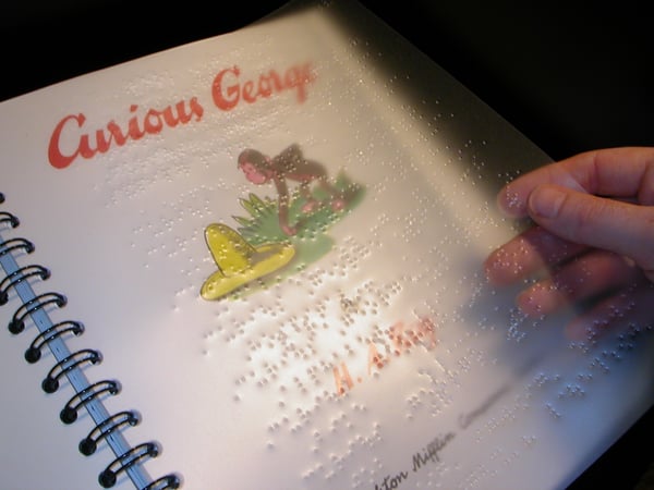 A copy of the original Curious George book with a plastic sheet of braille laid over its printed counterpart.